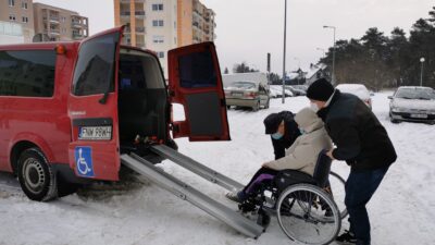 po lewej stronie fragment samochodu przystosowanego do transportu osób z niepełnosprawnością ruchową. Drzwi samochodu są otwarte, dwóch mężczyzn próbuje wciągnąć kobietę na wózku do samochodu. Jest zima, wszędzie leży śnieg. W oddali bloki mieszkalne, po prawej stronie las.