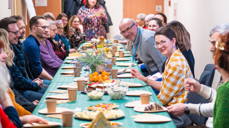 Na zdjęciu widać podłużny stół zastawiony różnymi potrawami. Przy stole siędzą uśmiechnięte osoby - kobiety i mężczyźni.