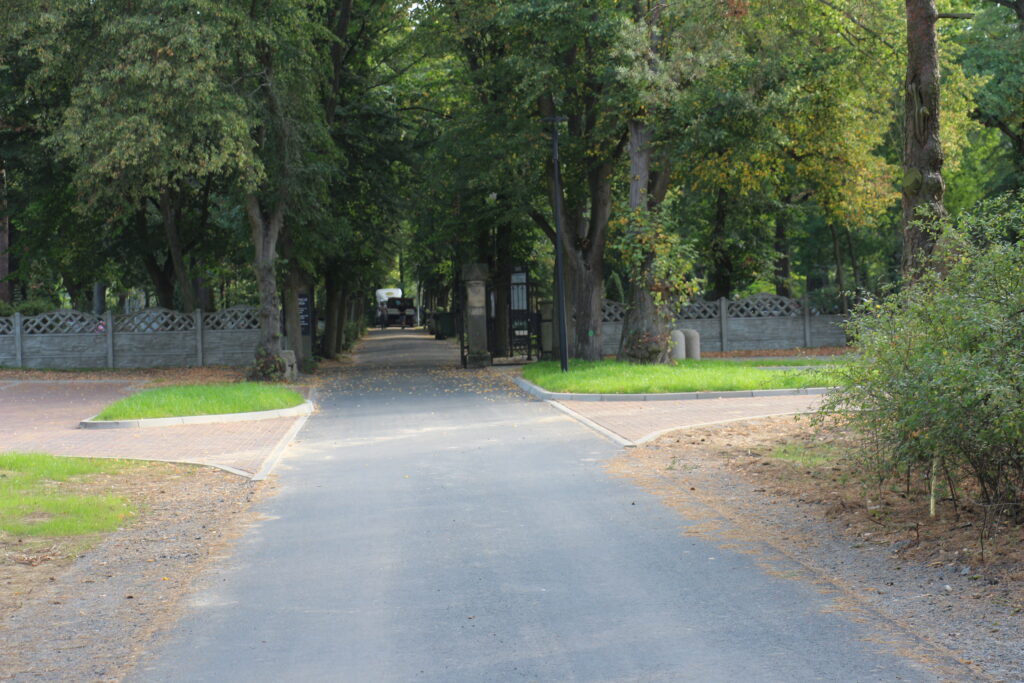 Asfaltowa droga do cmentarza przy ulicy Piaskowej w Nowej Soli w tle drzewa oraz brama wjazdowa
