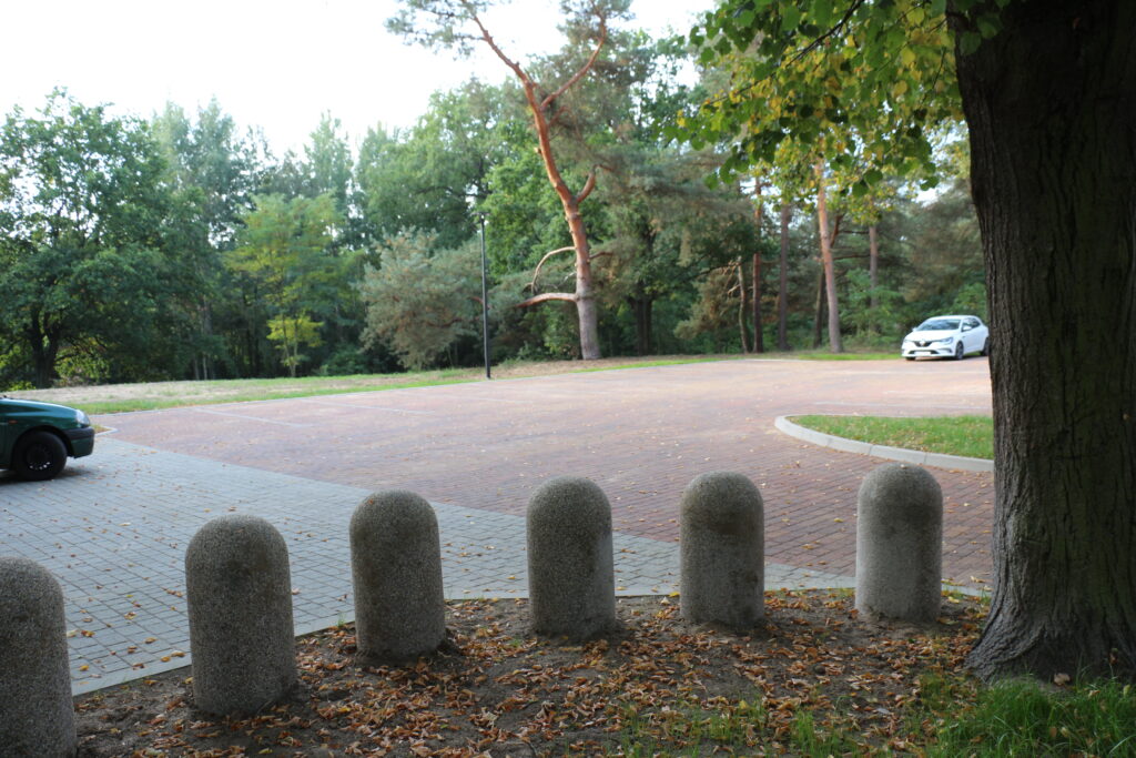 Parkingi przed wjazdem na cmentarz przy ulicy Piaskowej, na pierwszym planie kamienne słupki oddzielające część parkingów od drogi