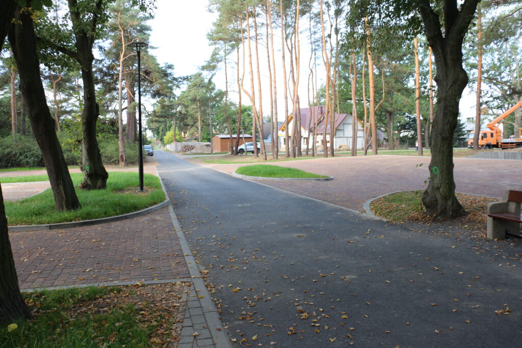 Widok na drogę doprowadzającą do cmentarza przy ulicy Piaskowej. Po obu stronach drogi parkingi przy których rosną drzewa. W tle domy i samochody