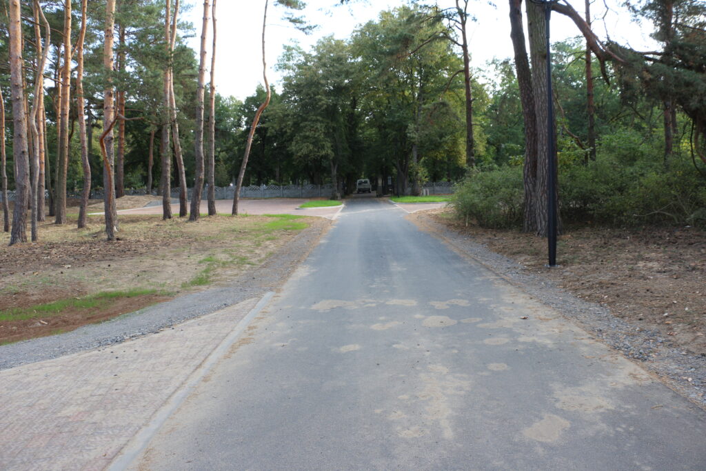 droga do cmentarza przy ulicy Piaskowej, po ubu stronach jezdni drzewa, w tle brama wjazdowa do cmentarza oraz jego ogrodzenie