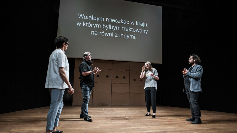 Na zdjęciu czworo aktorów Teatru Polska podczas spektaklu pod tytułem Jeden Gest. Stoją w kręgu i komunikują się za pomocą gestów. W tle ekran wyświetlający tekst ich rozmowy