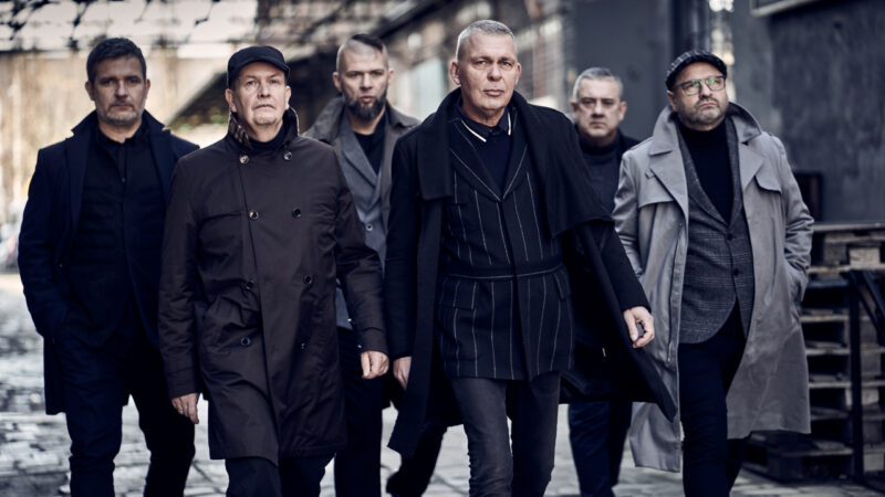 zdjęcie zespołu Raz Dwa Trzy. Sześciu mężczyzn w płaszczach, idą w budynku pofabrycznej hali