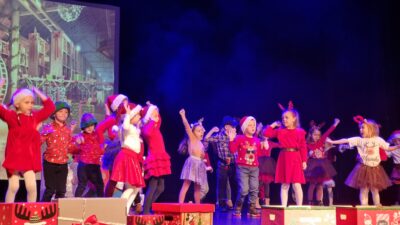 grupa dzieci w strojach świątecznych, uśmiecha się i tańczy, dzieci śpiewają, przed dziećmi na scenie pudełka prezentowe
