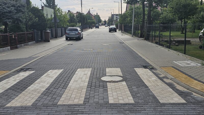 nowo wybudowana droga, po obu jej stronach nowe chodniki, w tle zaparkowane samochody, na pierwszym planie przejście dla pieszych